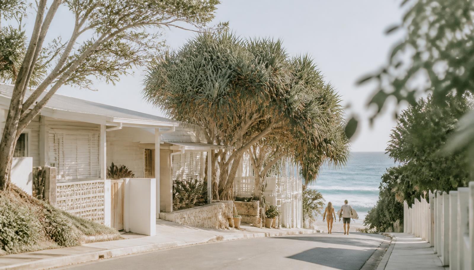 A sunlit beachside street of white coastal homes and pandanus palms leading down to the ocean